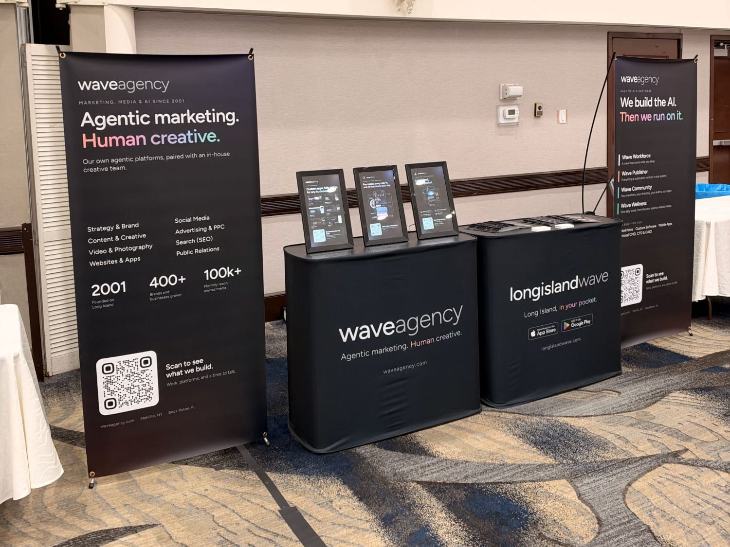 The Wave Agency booth at the Melville Chamber Business Expo: a banner reading Agentic marketing. Human creative., a Wave Agency table with three digital frames, a Long Island Wave table, and a second banner reading We build the AI. Then we run on it.