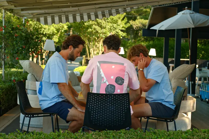 Friends at a garden table under a striped awning, one in a pink graphic tee.