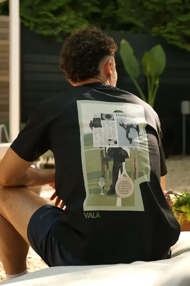 A man seen from behind in a black graphic tee, sitting in the shade.