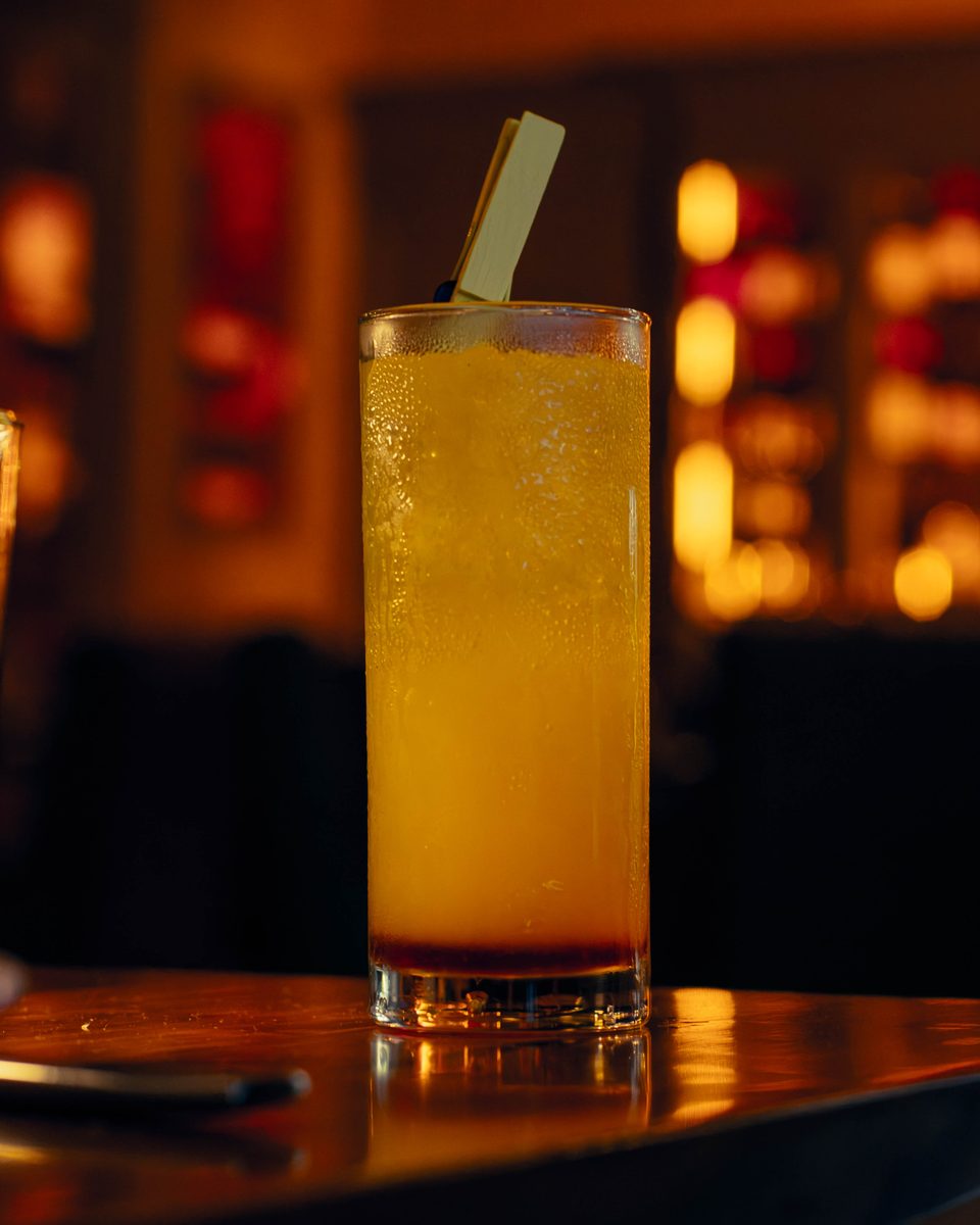 A tall citrus highball on the bar, the room warm and dark behind it.